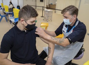 Firefighter giving a man a Covid 19 vaccination at a vaccination centre. They are both wearing face coverings.